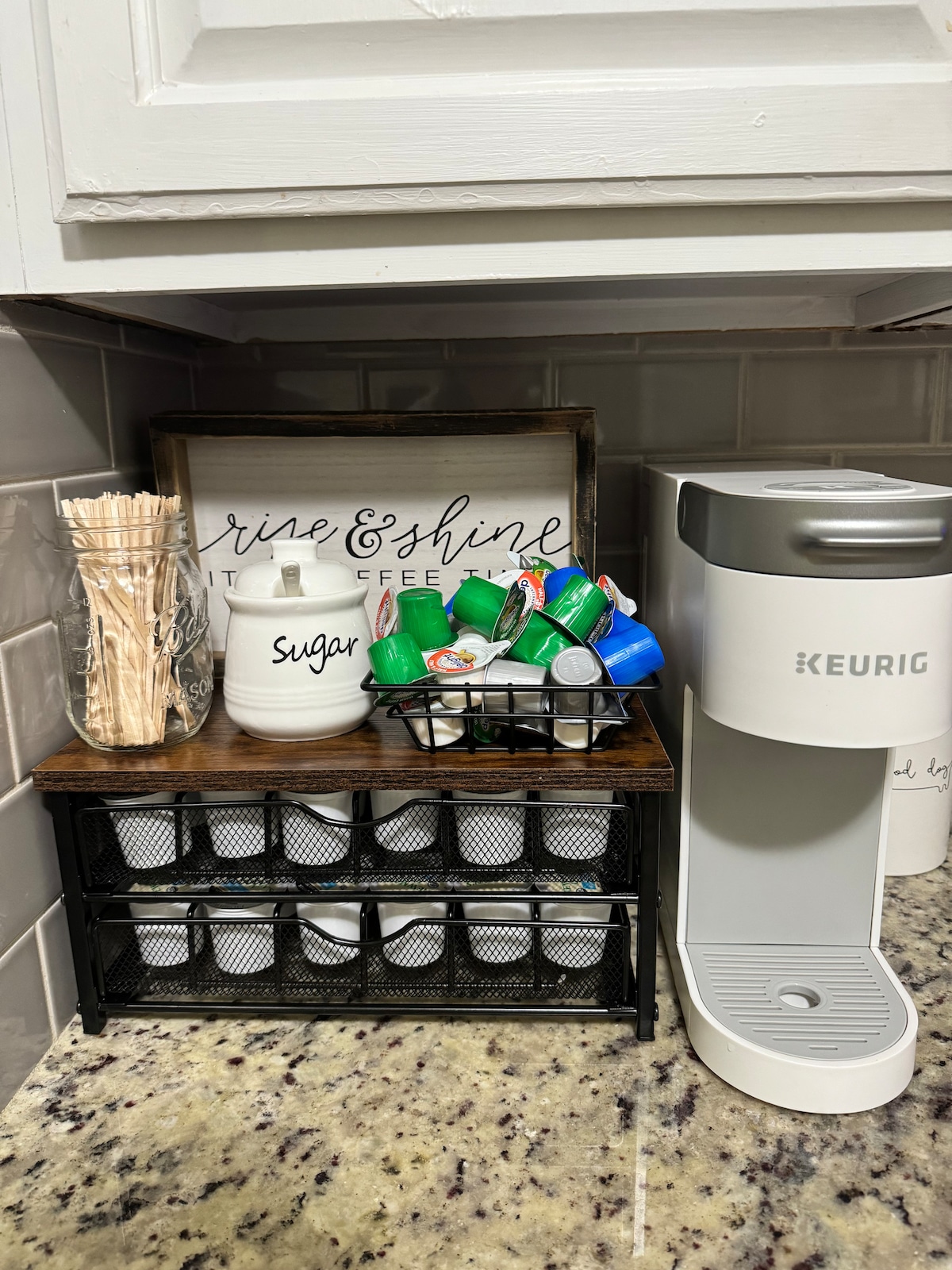 A neatly arranged kitchen counter features a Keurig coffee maker alongside a decorative sugar jar and a container of stirrers. A basket filled with colorful coffee pods is positioned below, providing easy access to various flavors.