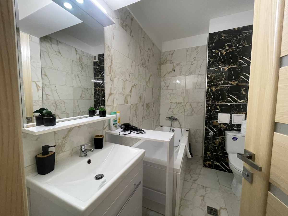 A modern bathroom is equipped with a white vanity featuring a sink and a mirror above. The walls are adorned with elegant marble tiles in light and dark hues, enhancing the space's contemporary design. A bathtub and a toilet are also visible, providing essential amenities.