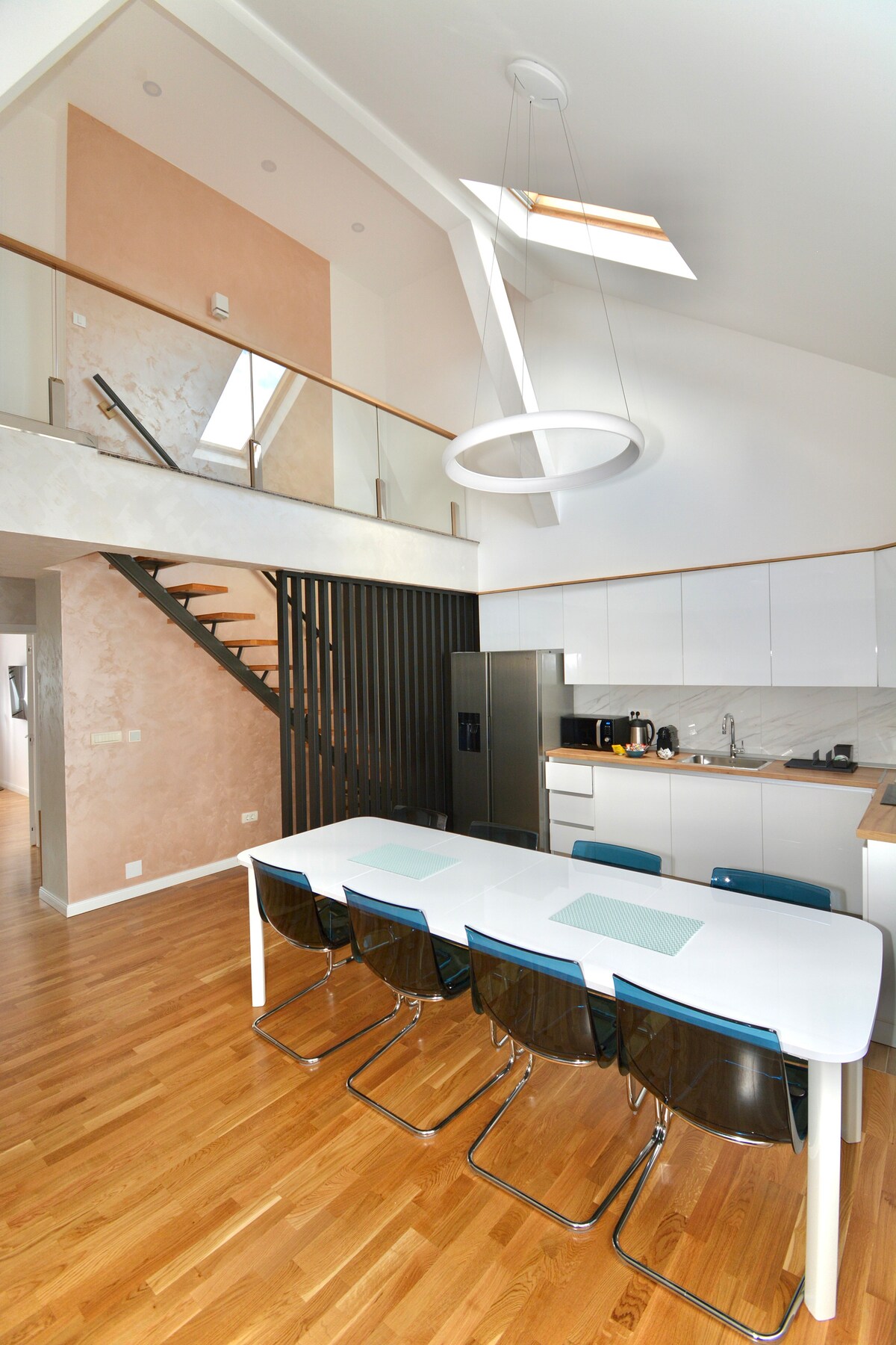 A spacious dining area is shown with a large white table surrounded by six transparent chairs. The modern kitchen features sleek cabinetry and stainless steel appliances. Natural light pours in from a skylight, illuminating the warm wood floors and highlighting the design elements of the duplex.