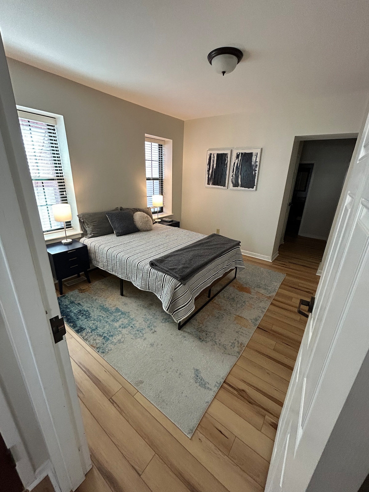 A well-lit bedroom is presented with a spacious bed dressed in soft linens. Two bedside lamps provide ambient light, while large windows reveal blinds. A subtle area rug is featured on the wooden floor, and framed artwork is mounted on the wall.