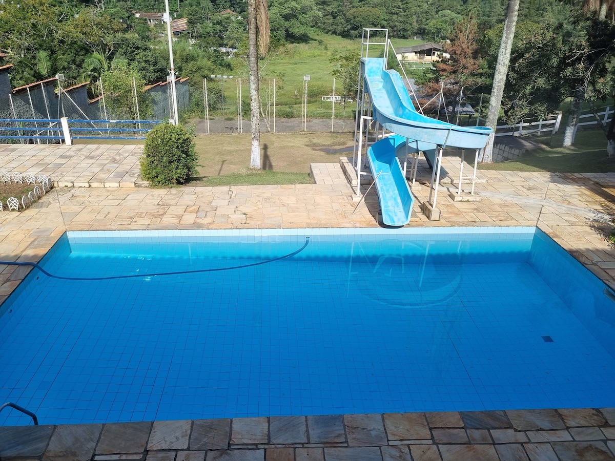 A swimming pool is featured with a slide on one side, surrounded by a tiled deck. Lush greenery and open space are visible in the background, enhancing the outdoor environment. The pool's clear water reflects the surroundings, creating a refreshing atmosphere.