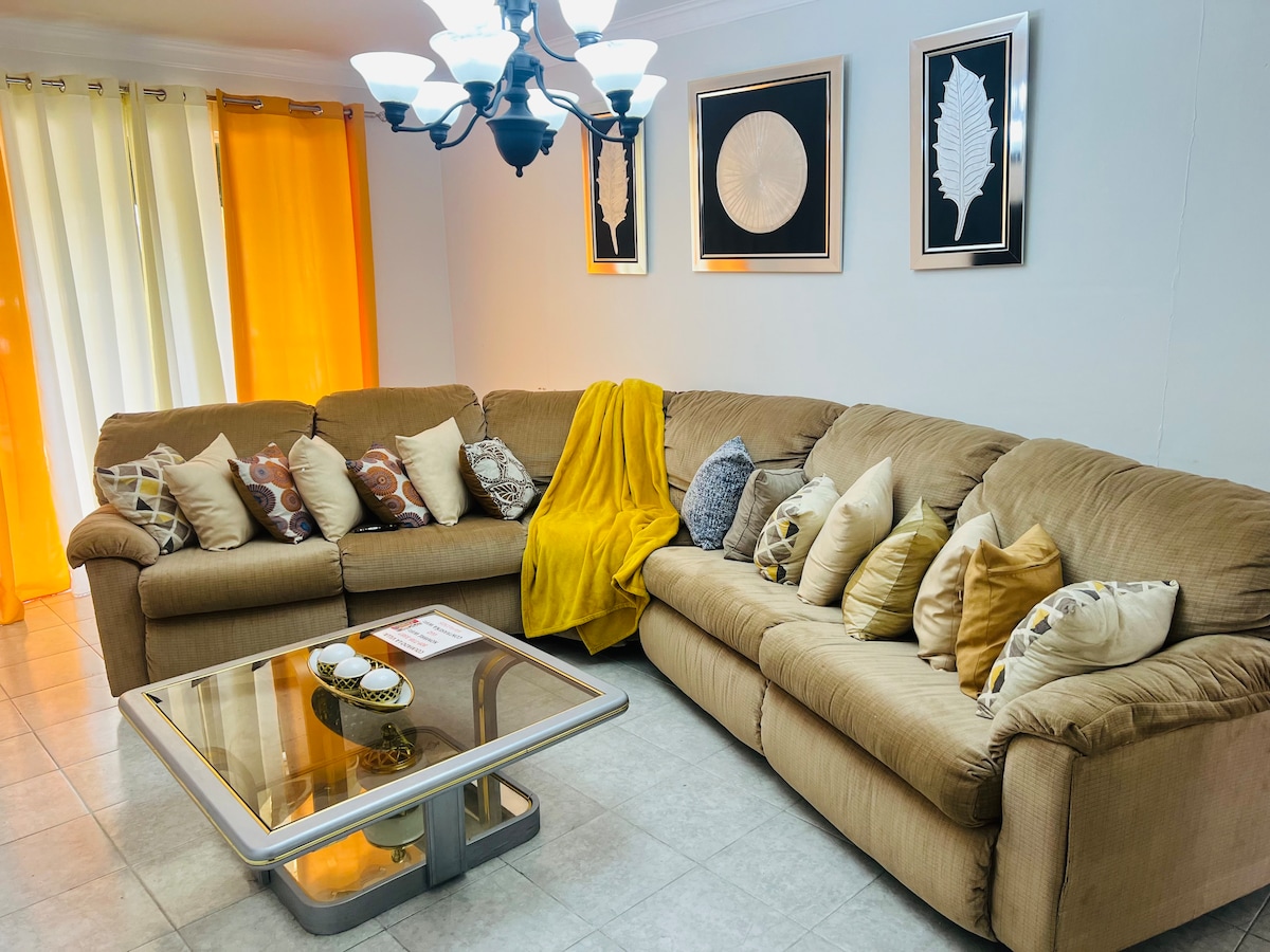 A spacious living area is featured, showcasing a large sectional sofa adorned with multiple decorative pillows. A coffee table is positioned centrally beneath a chandelier, while bright yellow curtains frame the windows, allowing natural light to brighten the room.