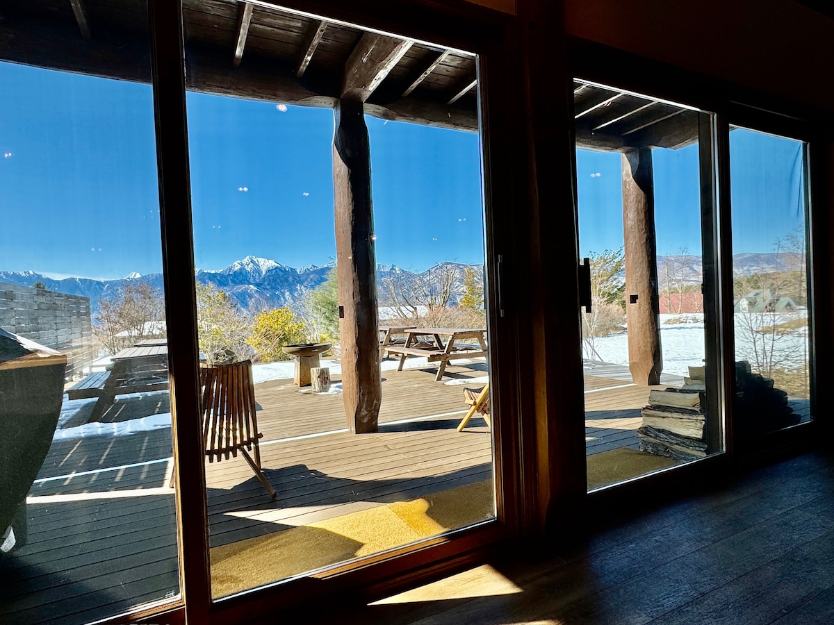 A spacious outdoor deck is visible through large glass doors, featuring wooden seating arrangements and mountain views. The landscape includes trees and a distant mountain range, with a clear blue sky above, creating a serene environment for relaxation.