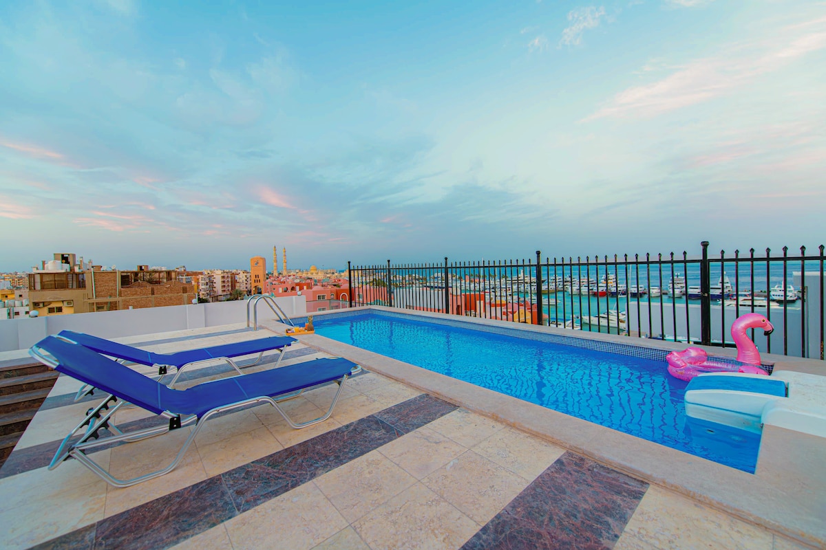 A rooftop swimming pool spans the terrace, surrounded by a black metal fence. Lounge chairs are positioned beside the pool, offering a view of the city and harbor. The sky features soft pastel hues, enhancing the setting's tranquil ambiance.