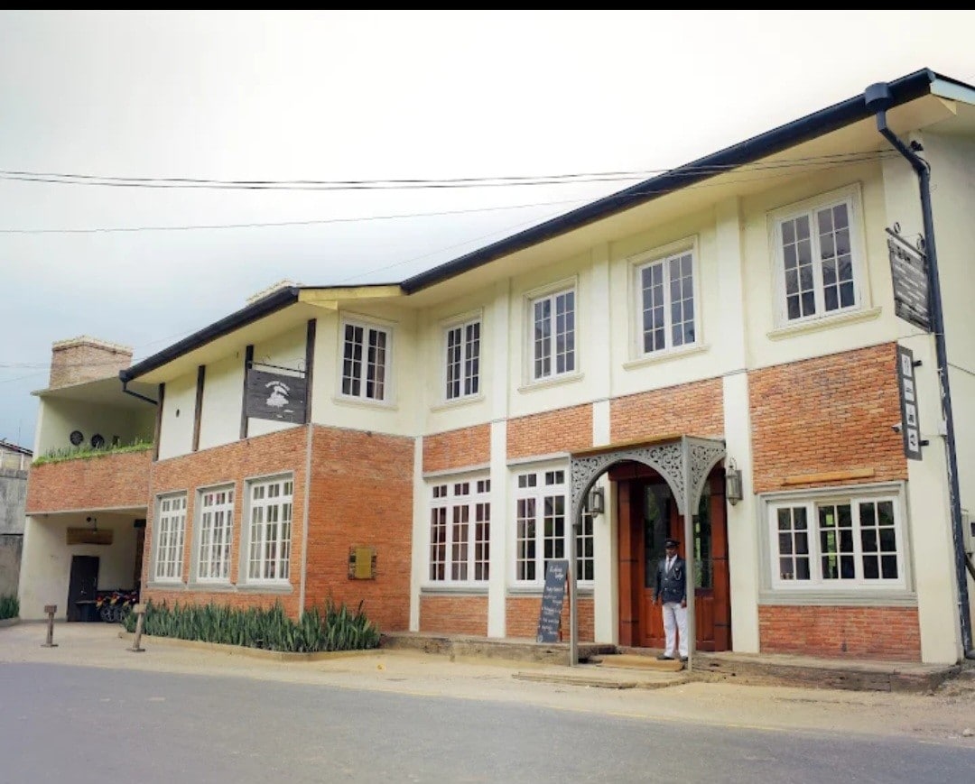 The exterior of the Railway Lodge Hostel showcases a charming colonial-style building. Large windows frame the red brick walls, while a decorative entrance is complemented by lush greenery along the pathway. The overall architecture reflects a warm and inviting design.