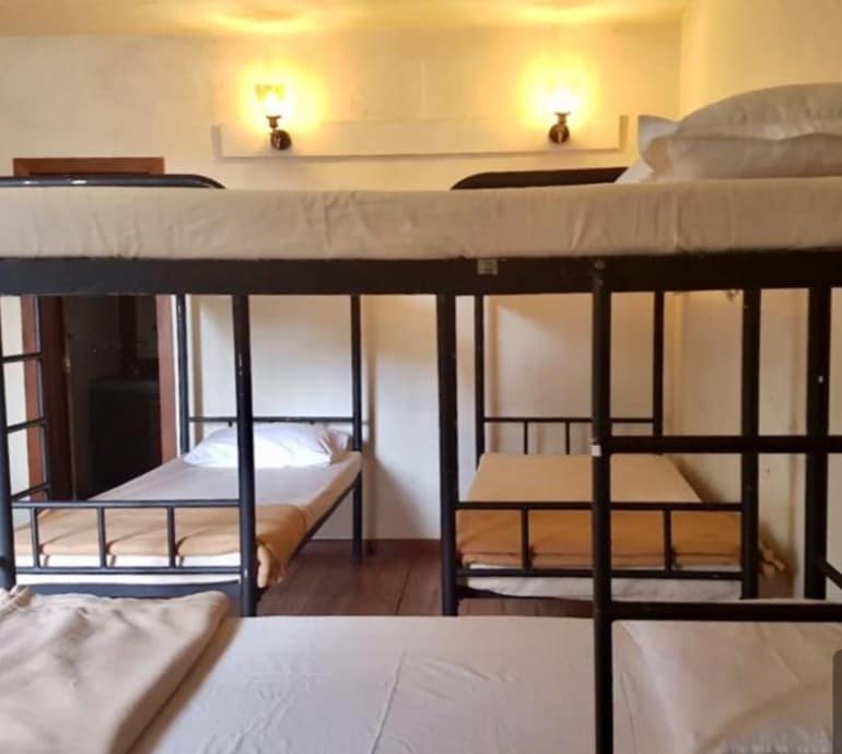 The image displays a dormitory with several bunk beds, providing ample sleeping space. Each bed features a simple mattress and bedding, arranged in a well-lit room. The soft glow from two wall sconces enhances the cozy environment, while the wooden floor adds warmth.