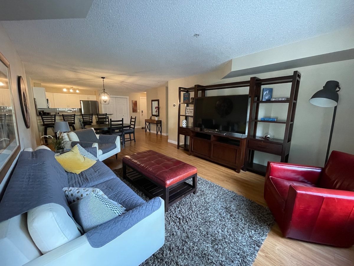 A spacious living area features comfortable seating, including a grey sectional and a red armchair. A coffee table sits atop a soft area rug, while a sleek entertainment unit holds a flat-screen TV. Dining chairs and a table are visible in the background, creating an inviting setting.