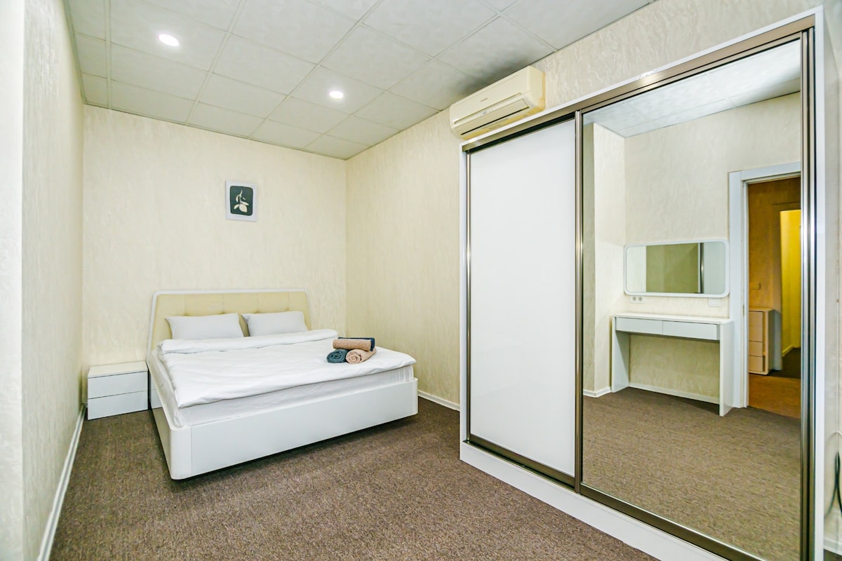 A light-colored bedroom features a spacious bed positioned against a wall. A large mirror and a vanity table are visible, alongside a closet with sliding doors. Air conditioning is mounted on the wall, and the floor is covered in soft carpeting.
