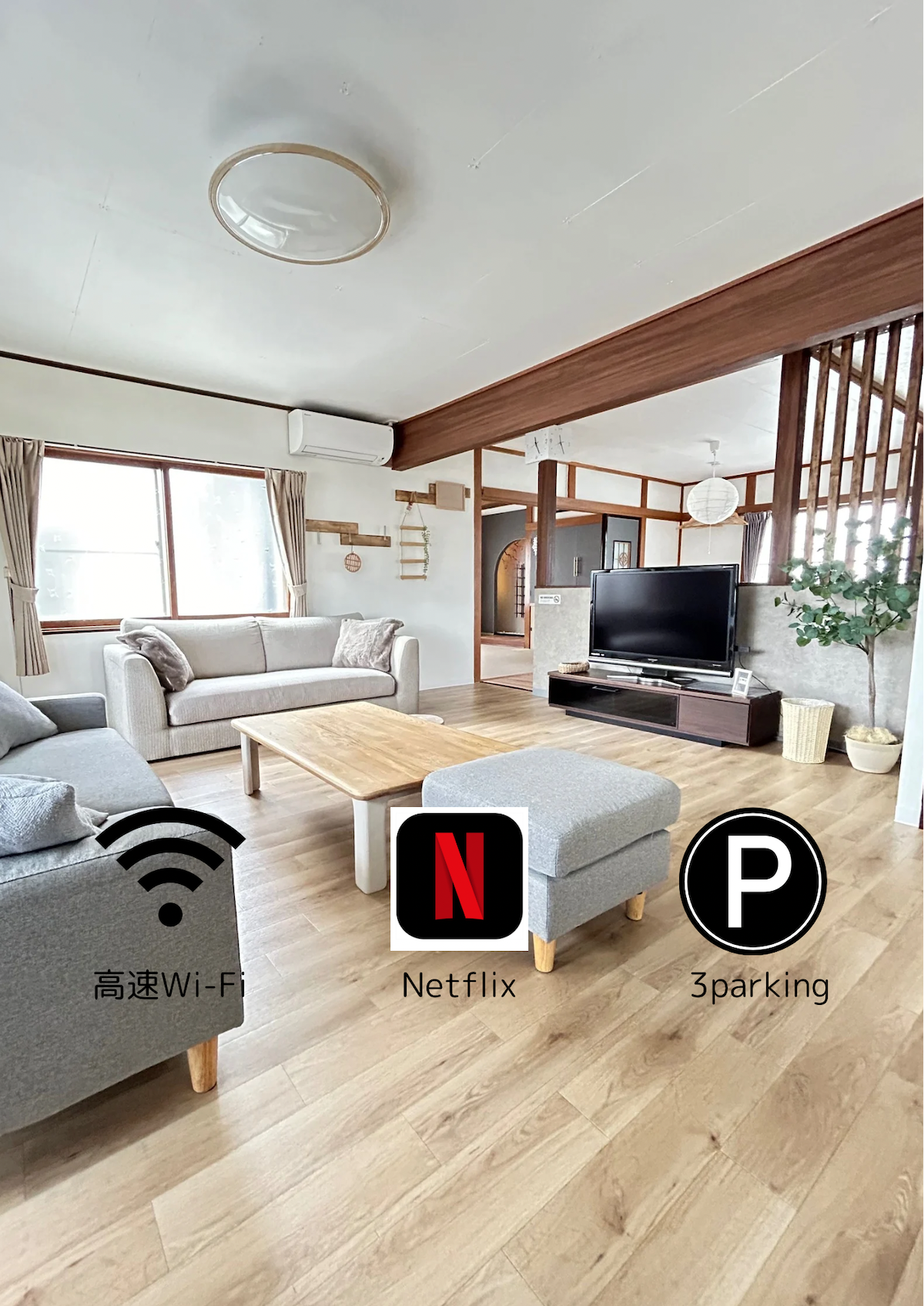 A spacious living room is furnished with two gray sofas and a wooden coffee table. Natural light fills the space through a large window, complementing the minimalist decor. A television is positioned on a stand, and icons for Wi-Fi, Netflix, and parking are visibly displayed.