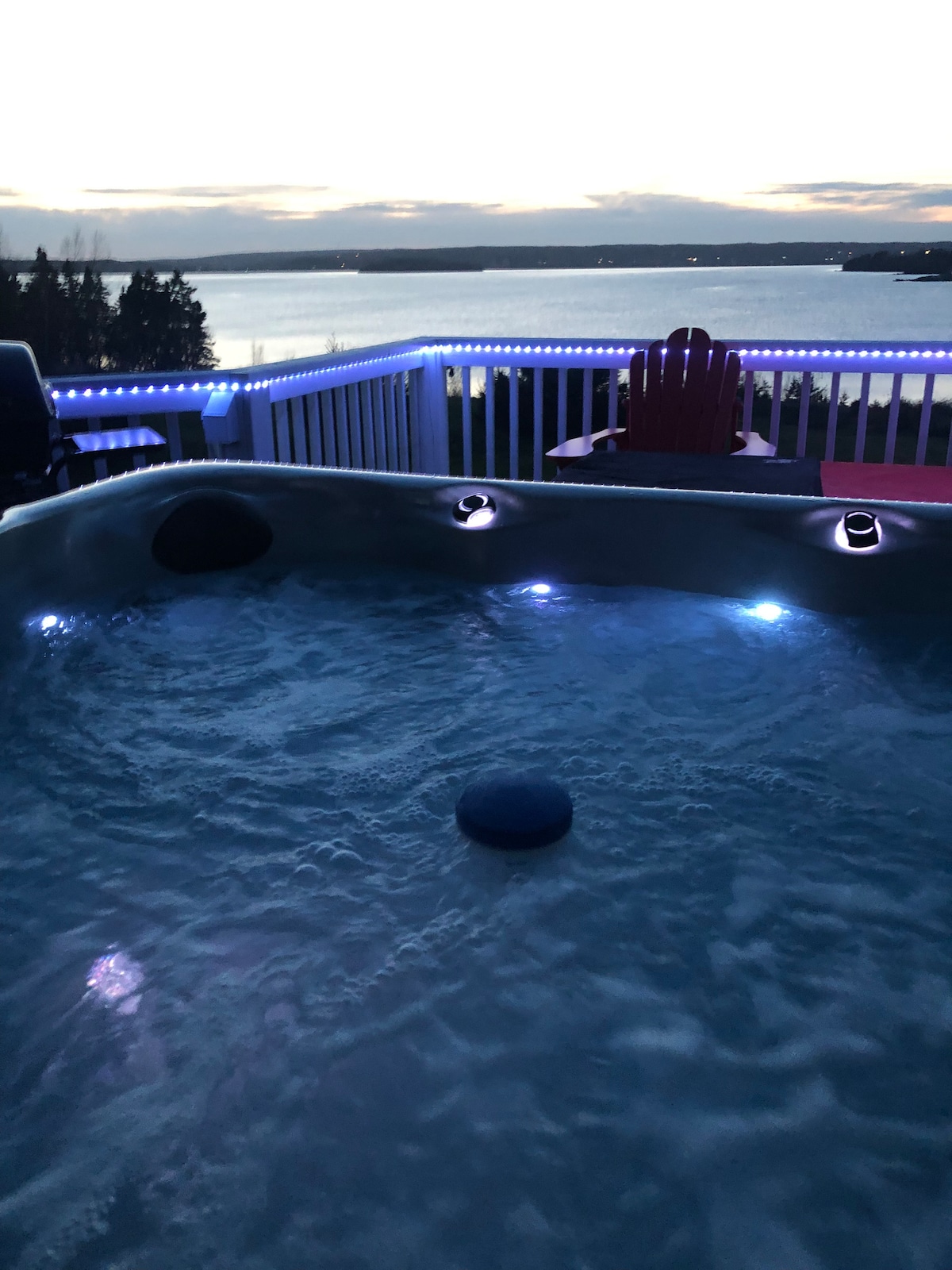 A private hot tub is illuminated with soft blue lights, inviting relaxation. The view captures the tranquil waters and the surrounding landscape, offering a serene atmosphere. The deck features a red Adirondack chair, enhancing the outdoor space for enjoying the scenic backdrop.