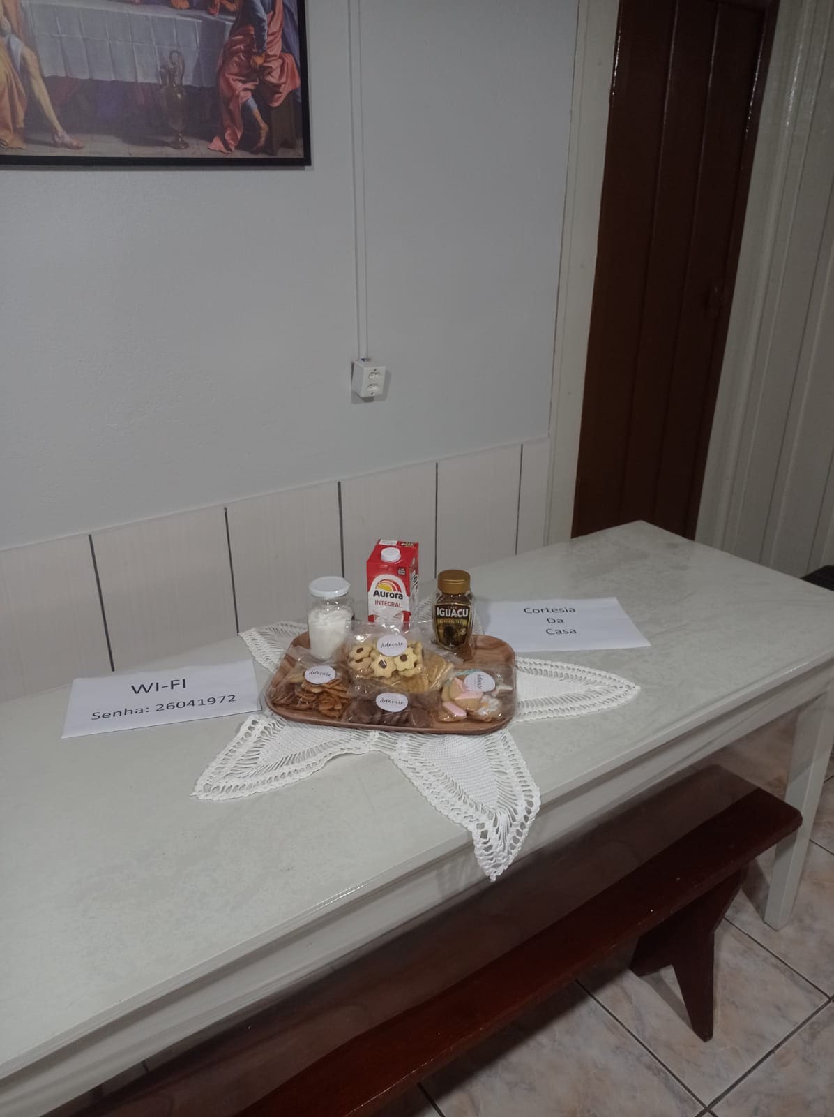 A dining table is set with a decorative woven placemat displaying a selection of snacks, including small cookies and a jar of chocolate spread. Nearby, printed cards provide information about Wi-Fi access. The space features light-colored walls and a wooden door leading to another area.