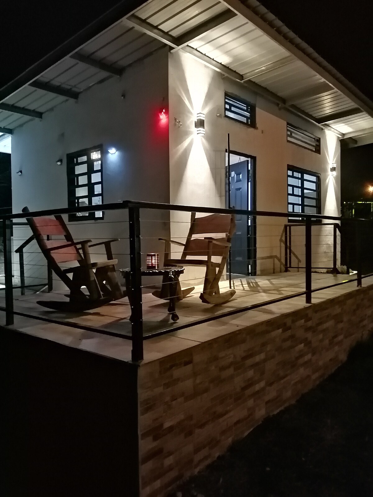 A cozy outdoor seating area is featured on the porch, with two rocking chairs positioned near a small table. Soft lighting illuminates the entrance of the tiny house, highlighting the modern design and creating a welcoming ambiance.