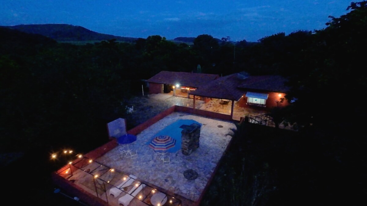 An aerial view captures a tranquil outdoor area featuring a stone patio, a refreshing pool, and a nearby terrace with lounge chairs. Surrounding nature creates a peaceful backdrop, while subtle lighting adds to the inviting atmosphere of the spacious setting.