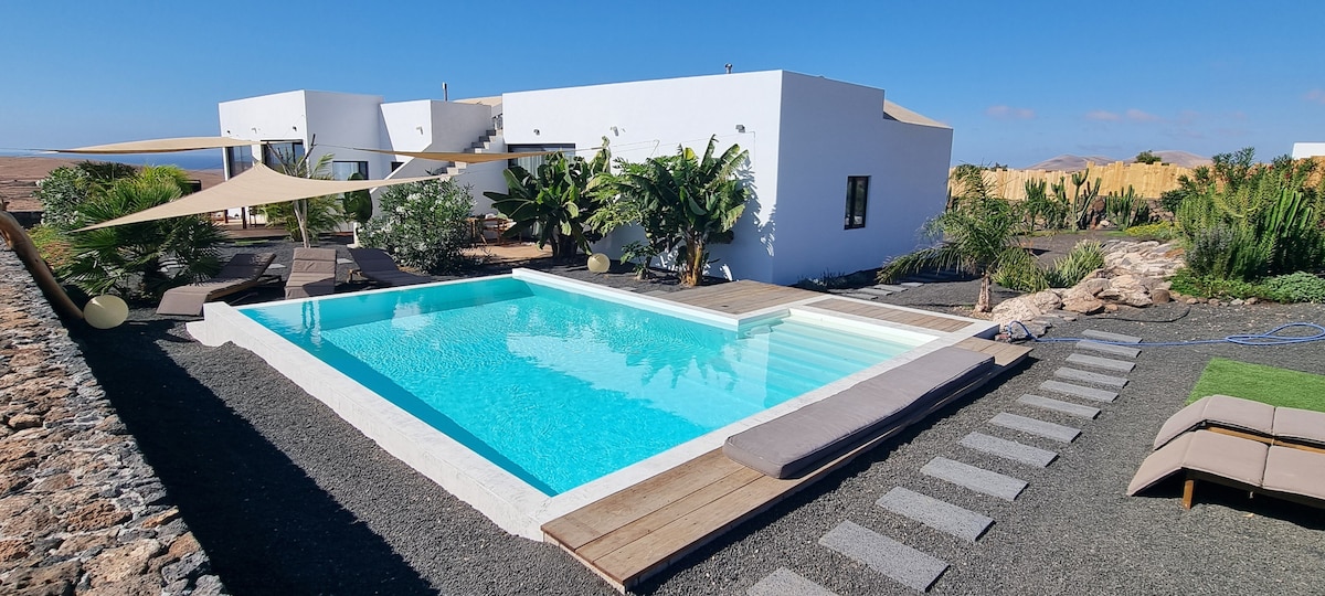 An inviting infinity pool reflects the clear blue sky, surrounded by lush greenery and tropical plants. Shaded lounging areas are positioned nearby on wooden decking, while a gravel path leads towards the main building, enhancing the serene outdoor setting.