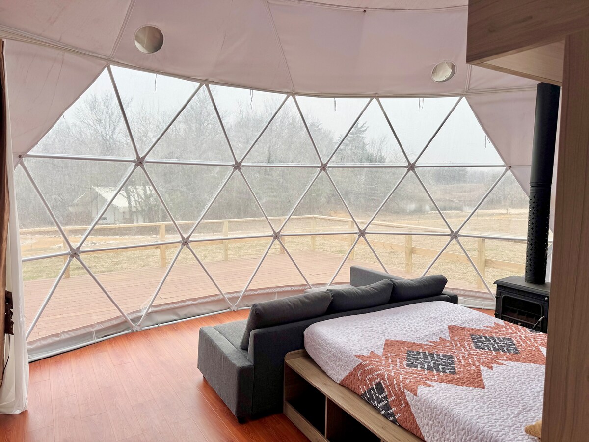 A spacious interior of a glamping dome tent features panoramic windows that provide a view of the surrounding landscape. A comfortable bed is positioned in the center, accompanied by a cozy seating area. Natural light fills the space, enhancing the serene atmosphere.