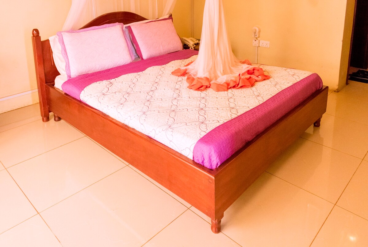 A spacious bed with a wooden frame features light-colored bedding accented with a vibrant purple coverlet. A decorative canopy hangs gently above, complementing the room's warm tones. The shiny tiled floor reflects the natural light entering from the adjoining area.