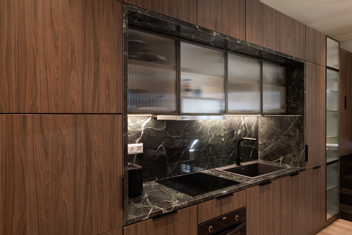 A modern kitchen features sleek brown cabinetry and a striking dark marble countertop. Built-in appliances are seamlessly integrated, including a modern oven and dual sink. Under-cabinet lighting highlights the elegant finishes, creating a polished and functional cooking space.