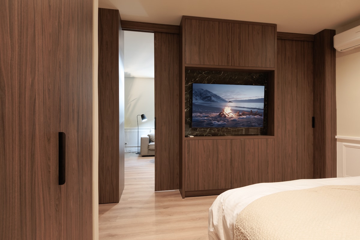 The image depicts a modern bedroom with wooden cabinetry, featuring a flat-screen TV mounted on the wall. The warm wooden tones are complemented by soft, neutral bedding. A doorway in the background leads into a stylish living area, enhancing the spacious feel of the layout.
