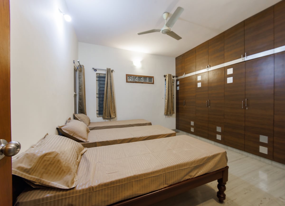A well-lit bedroom with two single beds positioned parallel to each other. Neutral-toned bedding complements the wooden bed frames. A large, stylish wardrobe with sliding doors occupies one wall, while curtains adorn the windows, allowing natural light to filter in.