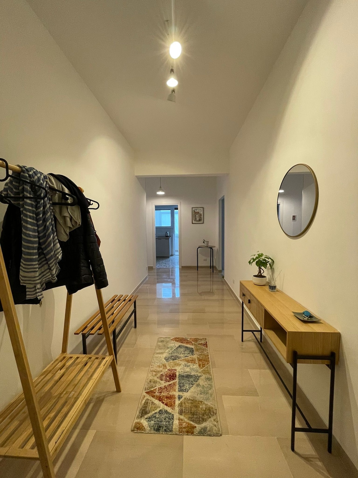 A well-lit hallway showcases a wooden rack for coats and hats alongside a sleek console table. A round mirror is mounted on the wall, reflecting light from overhead fixtures. A decorative rug adds color to the polished floor, leading towards an open door.