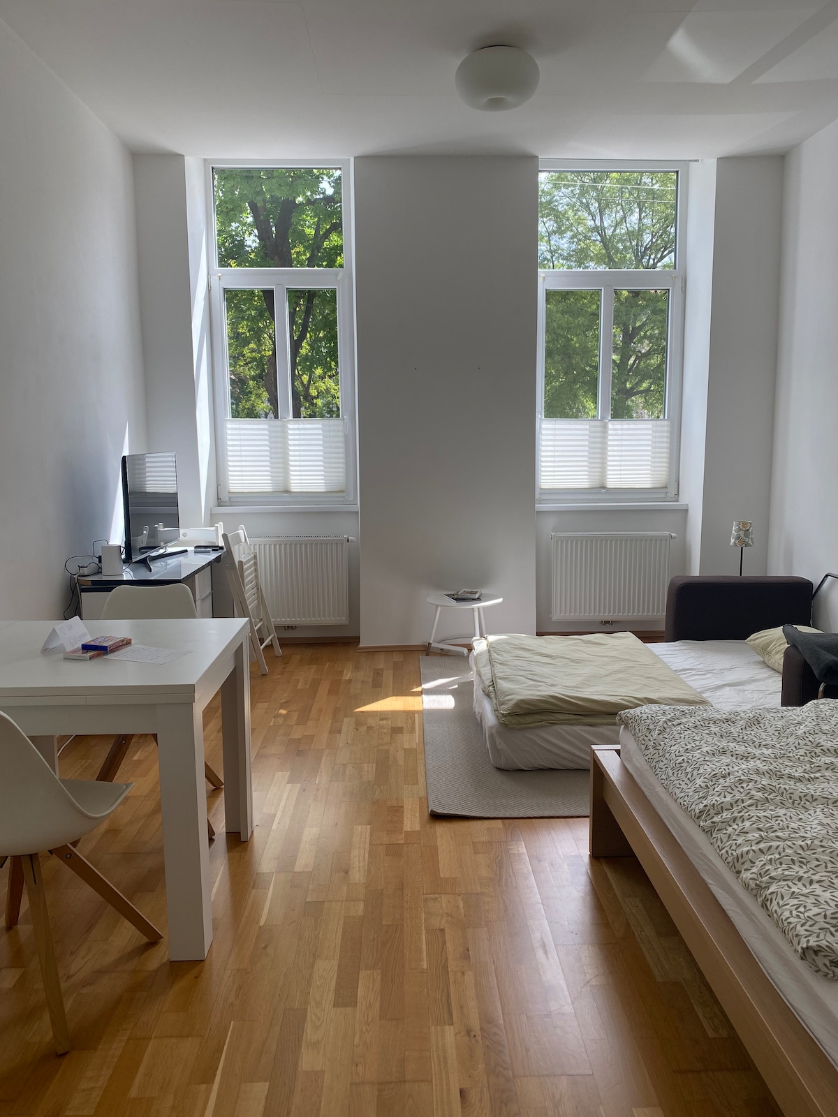 A bright room is illuminated by natural light streaming through two large windows. A spacious bed is placed against one wall, with a white table and chair situated nearby. A small armchair sits next to the bed, creating a functional living space.