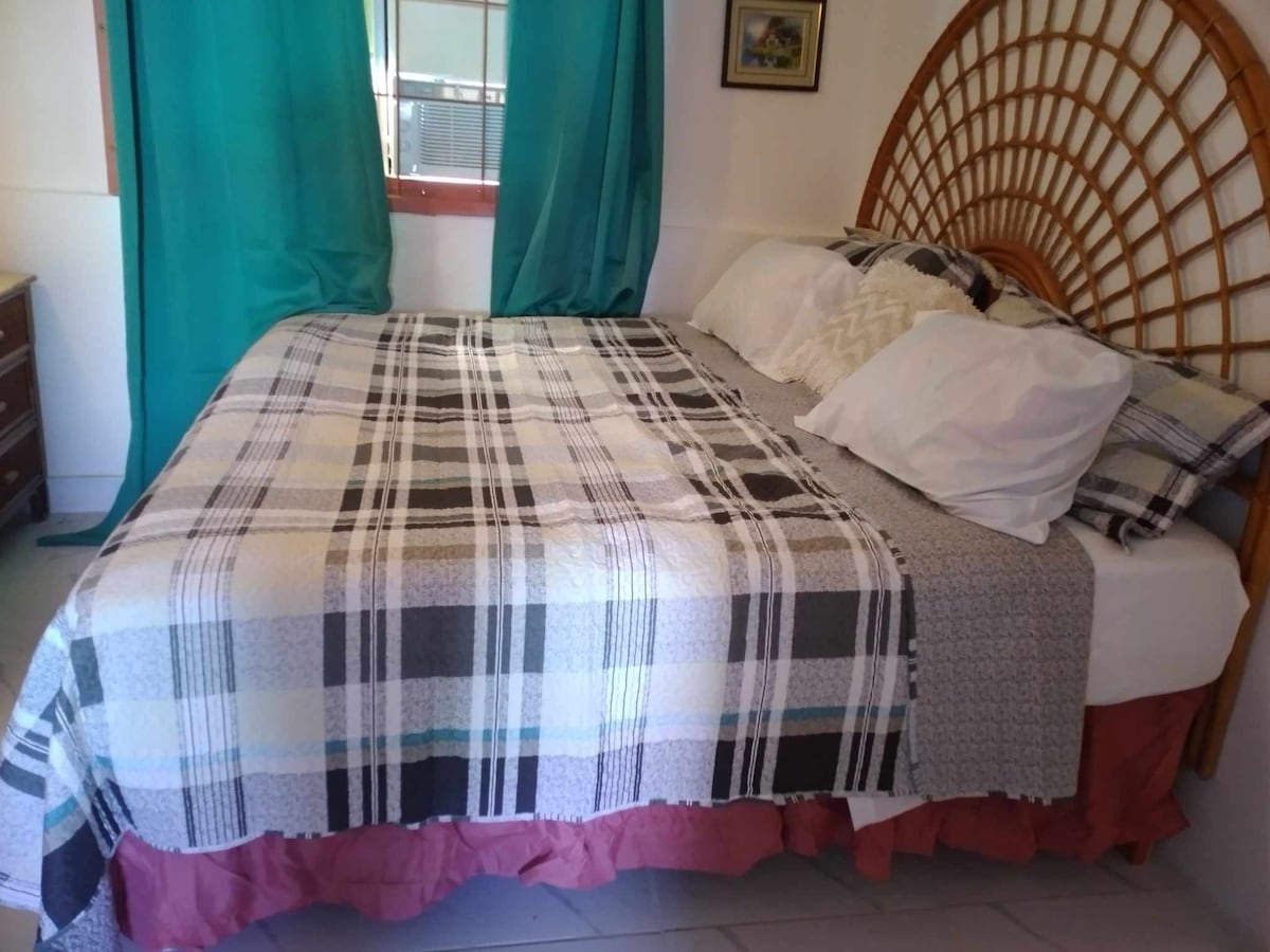 A cozy sleeping area features a spacious bed adorned with a patterned blanket. Two large pillows rest against a decorative headboard. Soft, turquoise curtains frame a window that allows natural light to brighten the room.