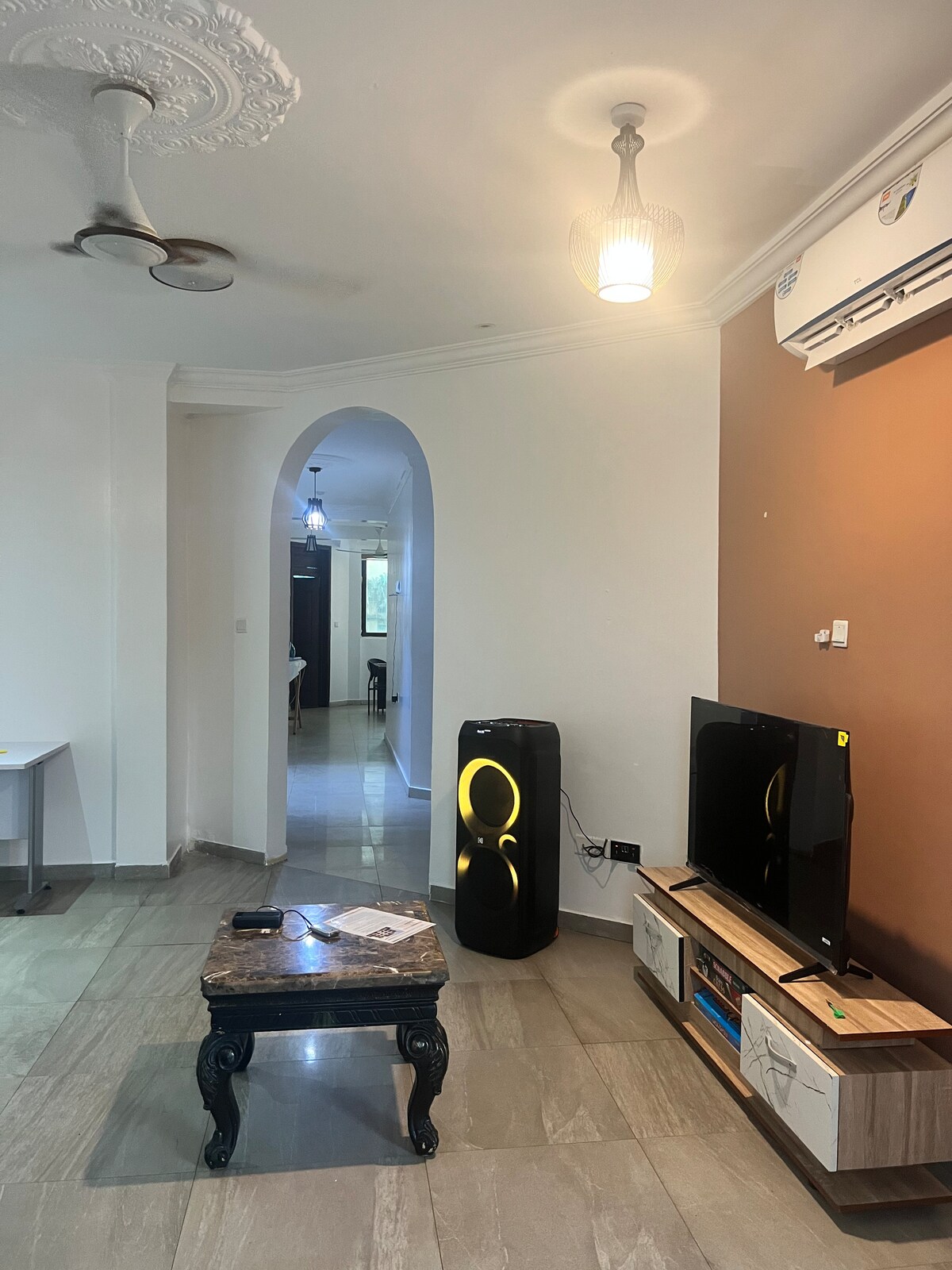 A living area is presented with a sleek television set atop a modern media unit. A stylish black coffee table occupies the center, while a floor lamp illuminates the room. An arched entryway leads into a hallway, adding architectural interest.