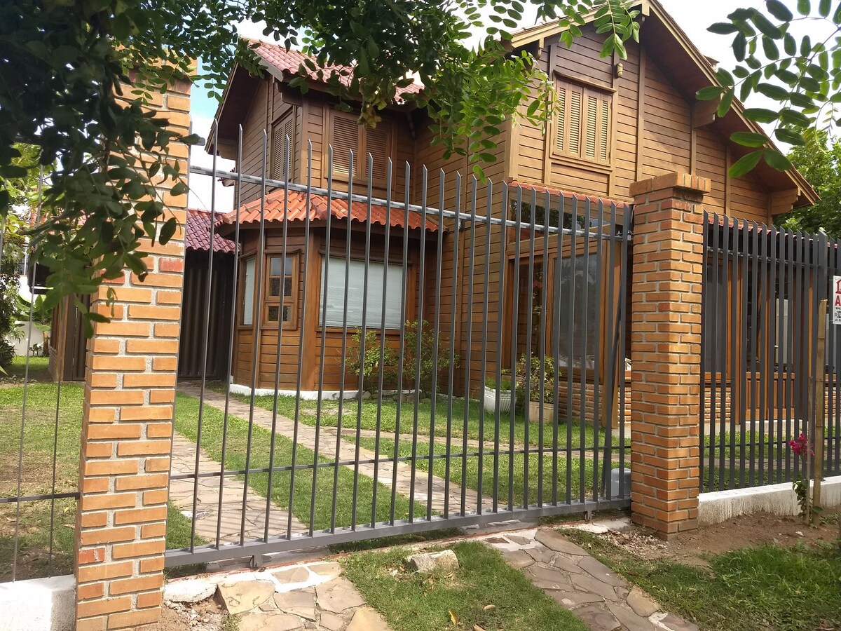 A charming wooden house is framed by a decorative metal fence, creating a welcoming entrance. The well-maintained garden features a stone pathway leading to the entryway. Large windows provide natural light, while greenery complements the warm tones of the exterior.