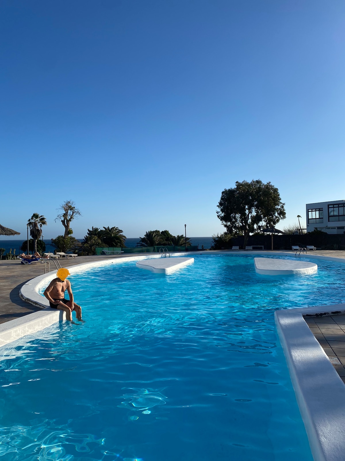 A community pool is visible with clear blue water. The pool features a gently curving shape and is framed by a tiled deck. Lush greenery and palm trees surround the area, while the distant ocean can be seen under a clear blue sky.