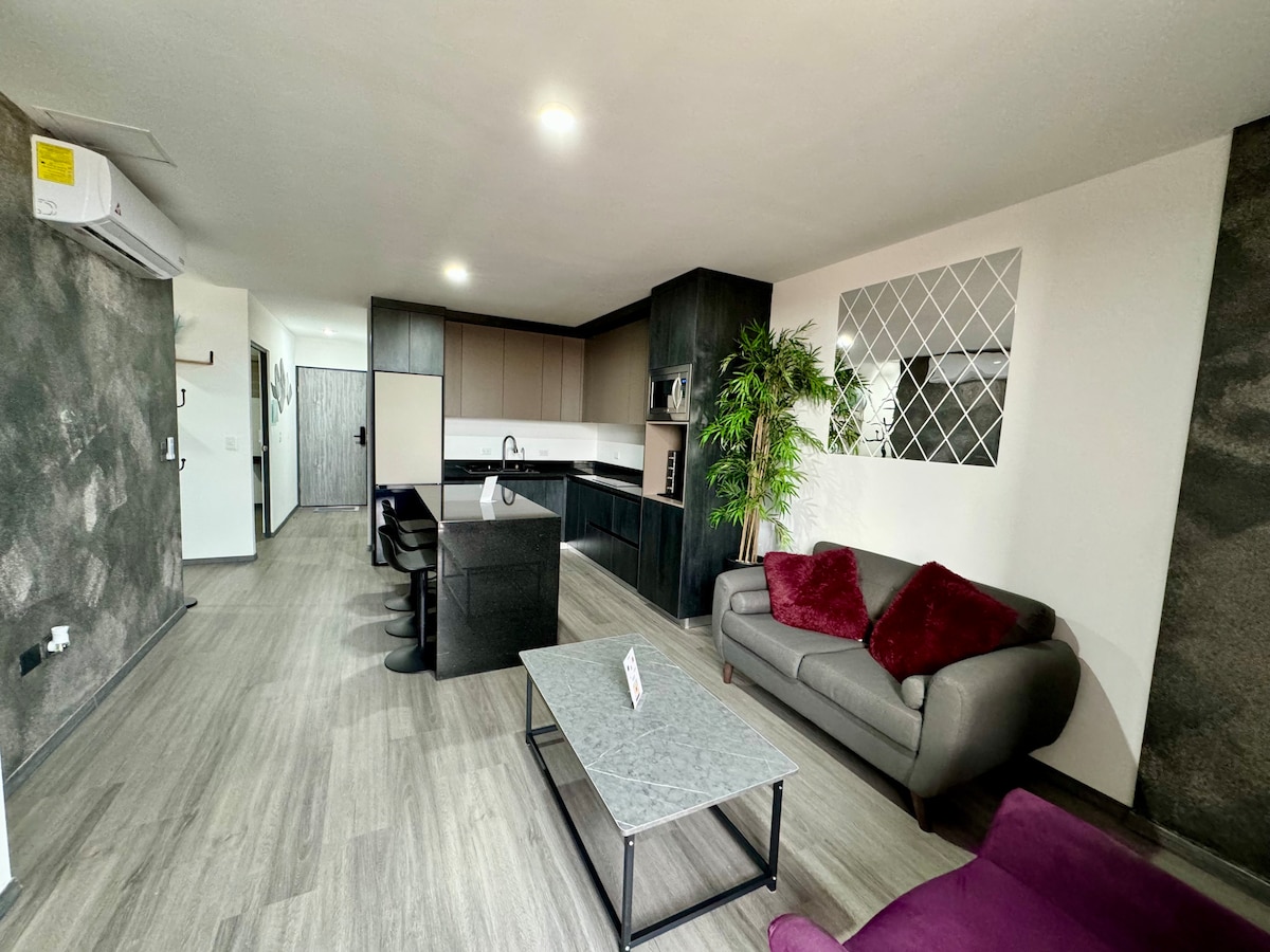An open-plan living area features a sleek kitchen with modern cabinetry and a central island. A comfortable gray sofa is complemented by burgundy accent pillows, with a minimalist coffee table positioned in front. A decorative wall with greenery adds a touch of elegance.
