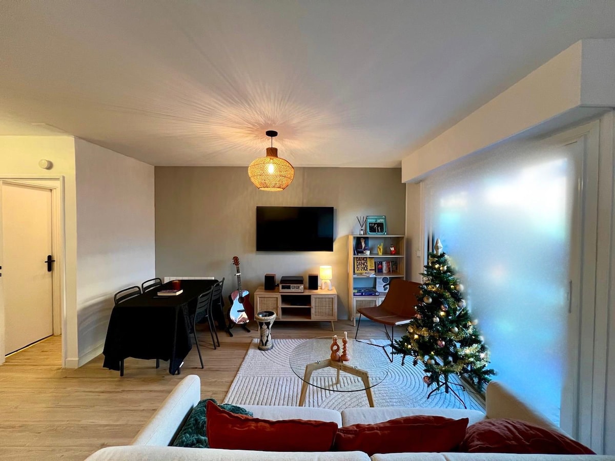 A living area is presented with a neutral color palette, featuring a soft sofa and a decorative rug. A coffee table is positioned in front of a television mounted on the wall. A small dining table is set for four, complemented by a Christmas tree and a cozy accent chair.