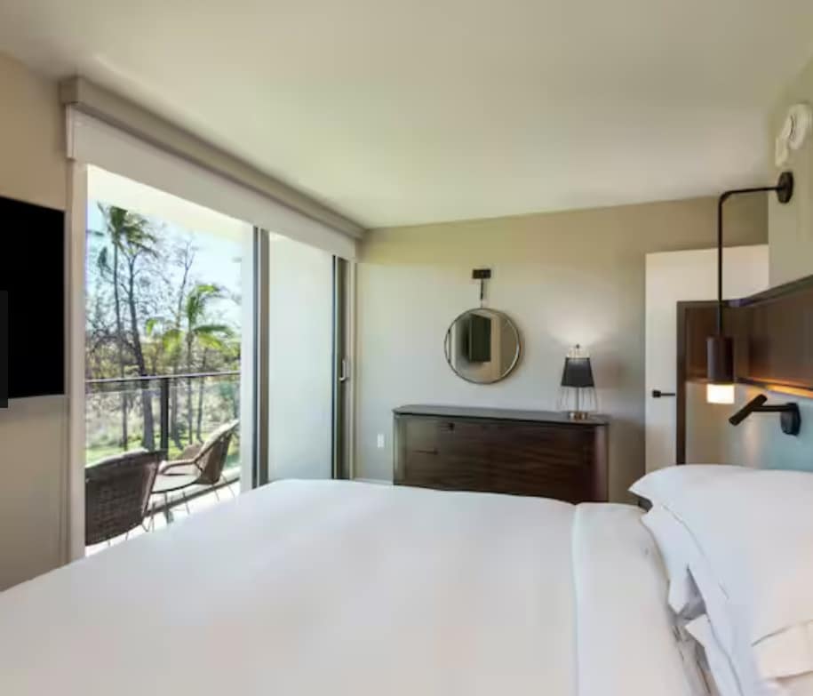 A bright bedroom features large sliding glass doors leading to a private balcony. A spacious bed is positioned prominently with crisp white linens, while a modern dresser and a round mirror complement the décor. Natural light enhances the inviting atmosphere.