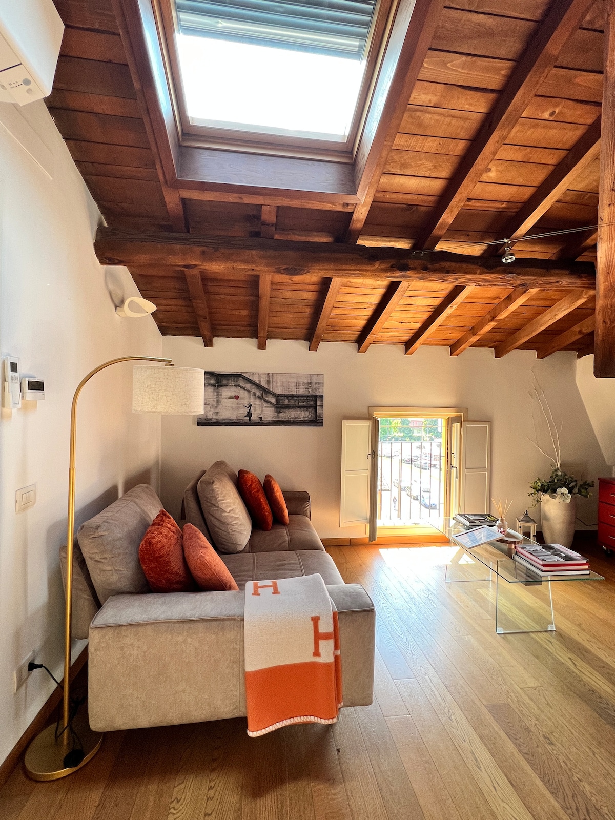A living area is presented with natural light streaming through a skylight. A comfortable sofa with orange accent pillows is positioned towards a window with shutters, while a sleek coffee table and minimalist artwork adorn the space. Exposed wooden beams highlight the ceiling's design.