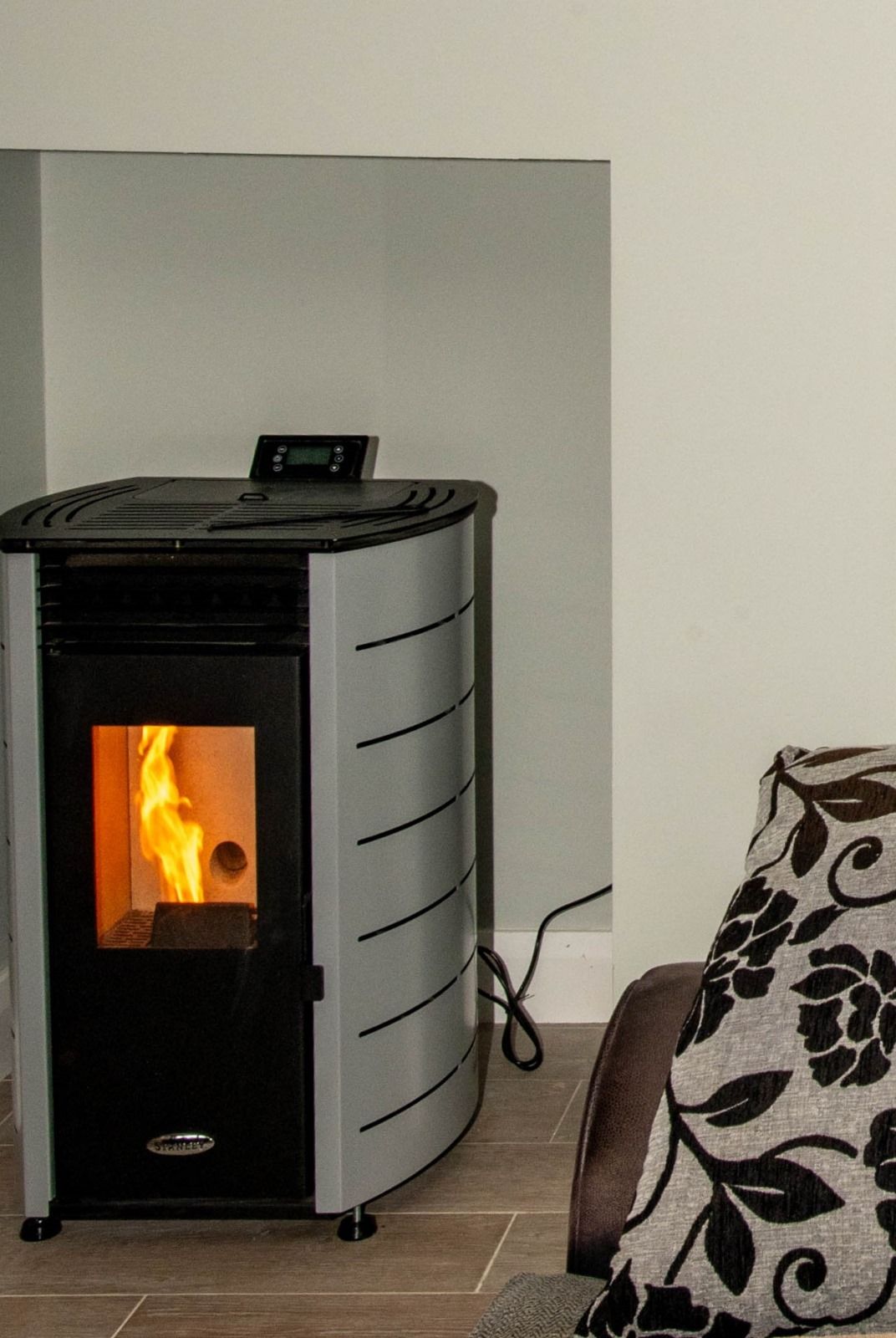 A modern heating unit is situated in a corner, featuring a large glass front displaying a gentle flame. The unit's sleek design is complemented by a neutral wall backdrop. A portion of a comfortable sofa, adorned with a floral-patterned cushion, is visible in the foreground.
