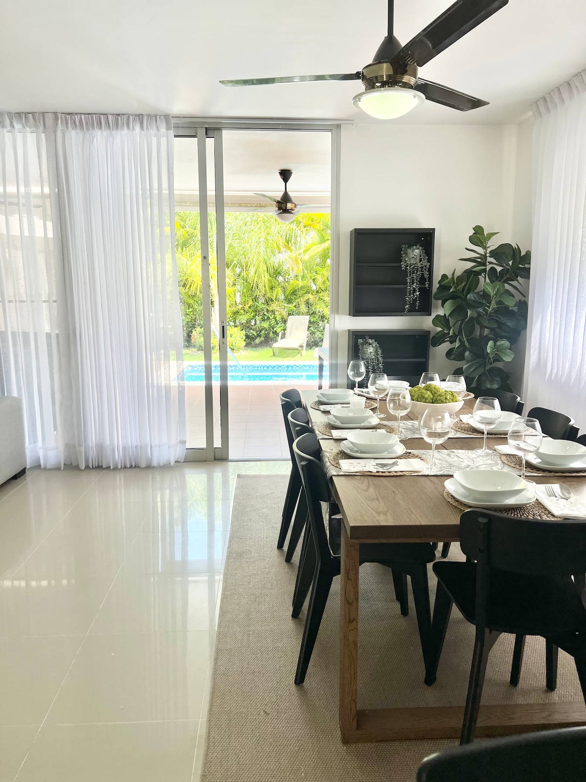 A dining area features a large wooden table set for eight, complemented by black chairs. Natural light enters through expansive glass doors, revealing a private pool area lined with lush greenery. Light-colored curtains gently frame the windows, adding a serene touch to the space.