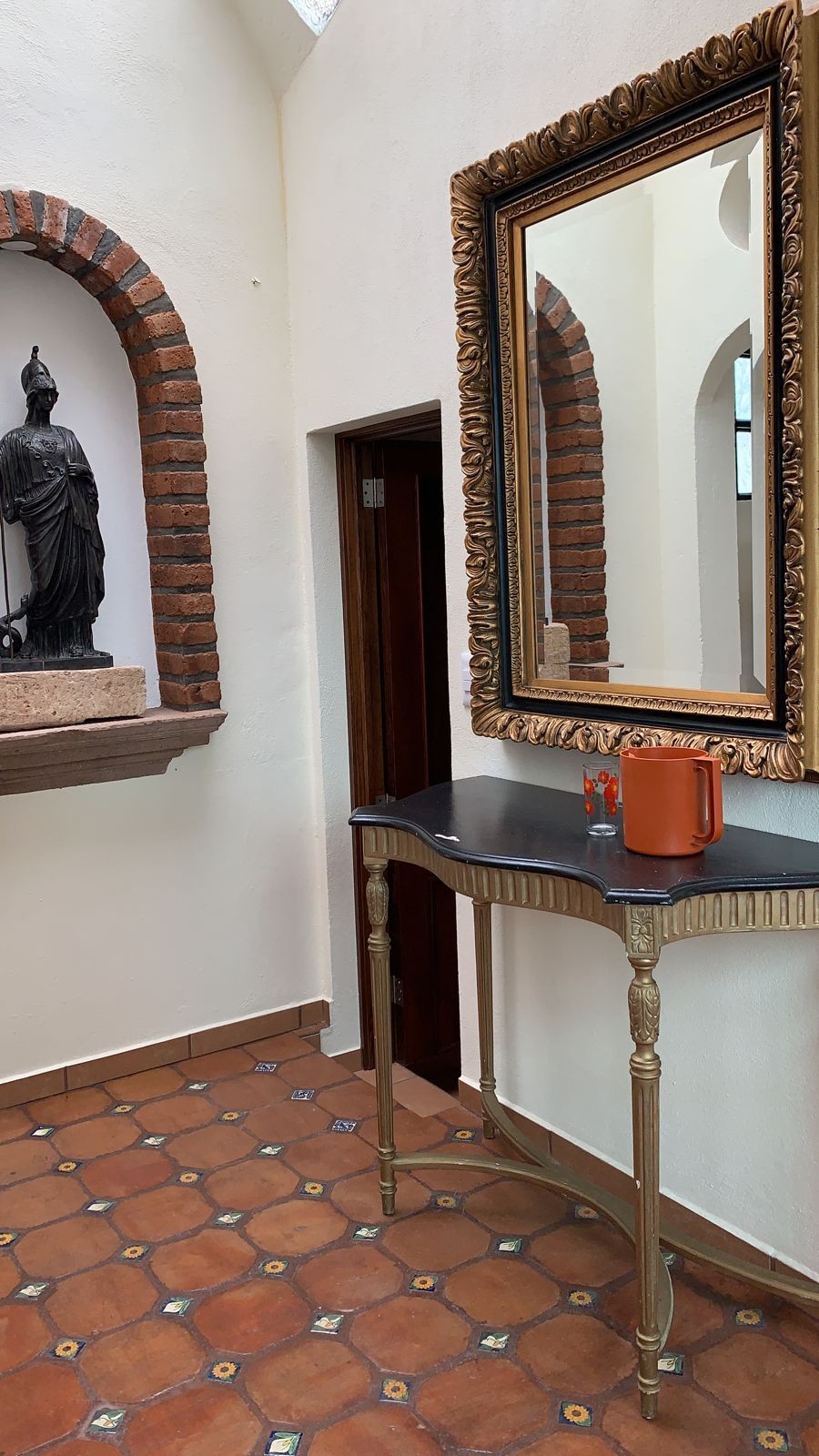 An elegantly styled entryway features a large ornate mirror above a sleek black console table. A terracotta tiled floor complements the warm tones of the brick archways and walls. A decorative statue is displayed in an alcove to the left, enhancing the space's historic charm.