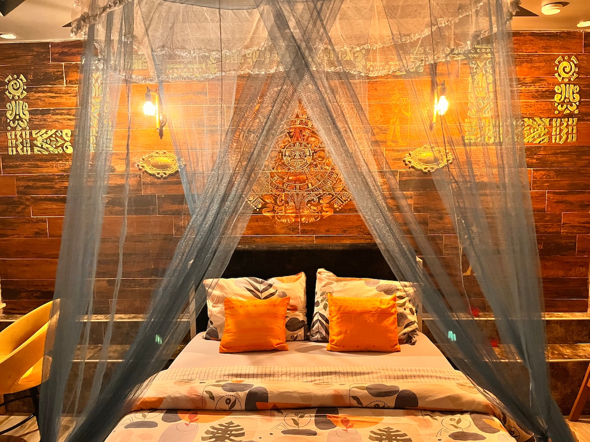 A sleeping area is presented with a comfortable bed, framed by a decorative canopy. The wall behind features intricate designs, complemented by soft amber lighting. Two vibrant orange cushions are placed on the bed, enhancing the cozy ambiance of the space.