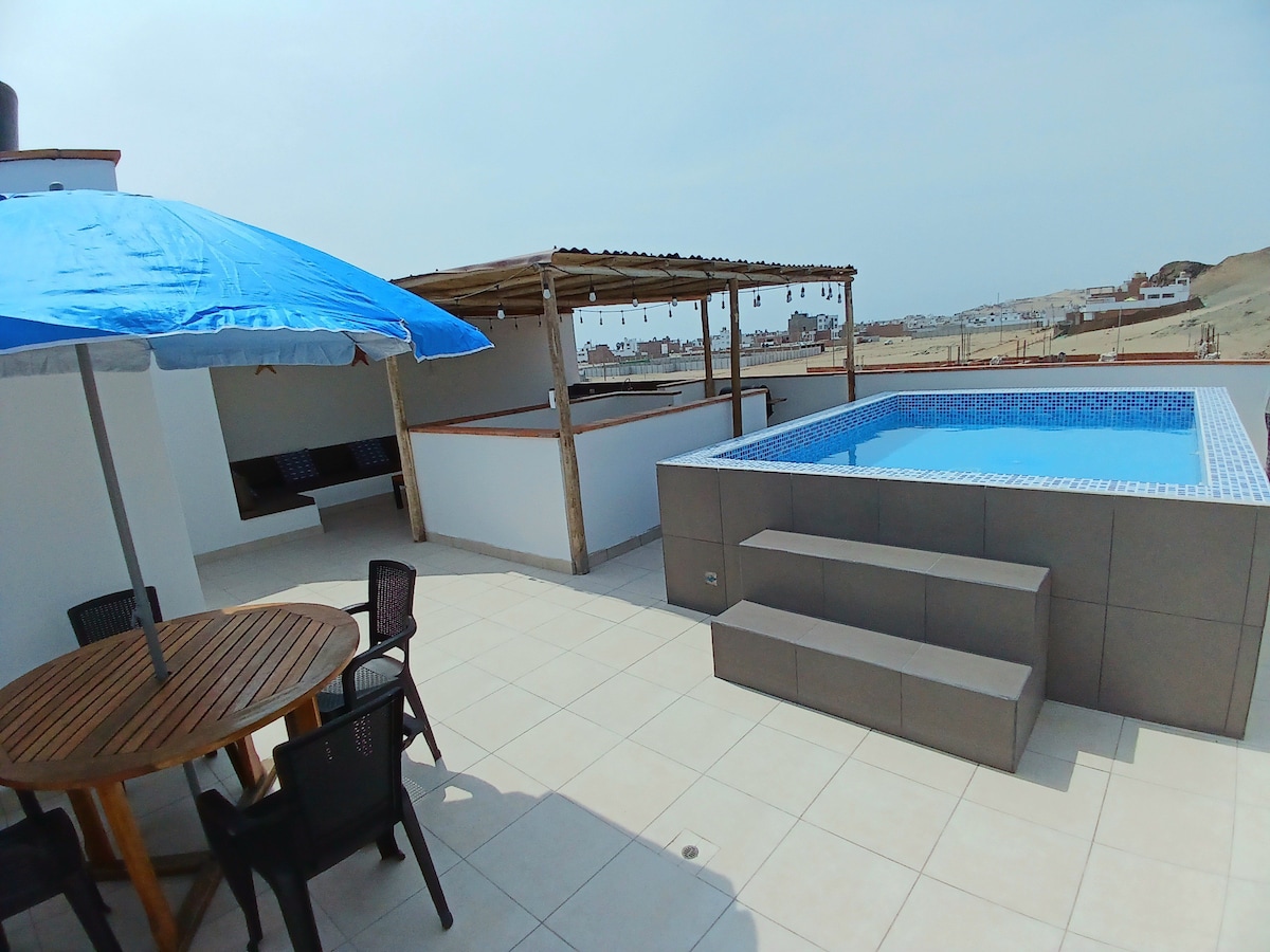 An outdoor terrace features a pool with steps leading in, surrounded by a shaded area with seating. A wooden table with chairs is positioned nearby, providing a suitable space for dining or relaxation. The terrace offers views of the surrounding landscape.