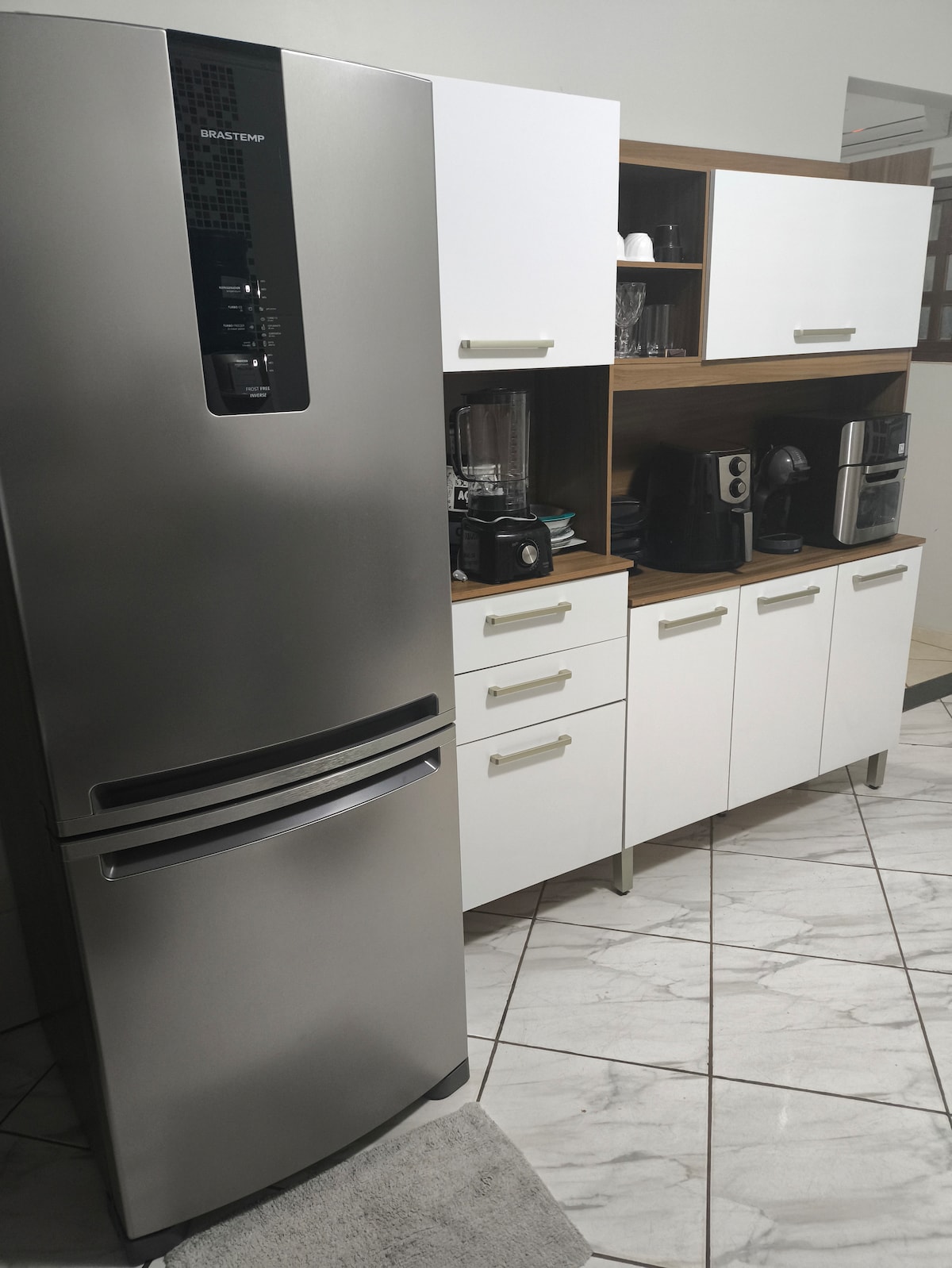 A modern kitchen area is featured, showcasing a stainless steel refrigerator alongside a white cabinetry unit. The cabinetry holds a coffee maker and other appliances, contributing to a functional and organized cooking space. A neutral-colored rug is positioned in front of the cabinetry.
