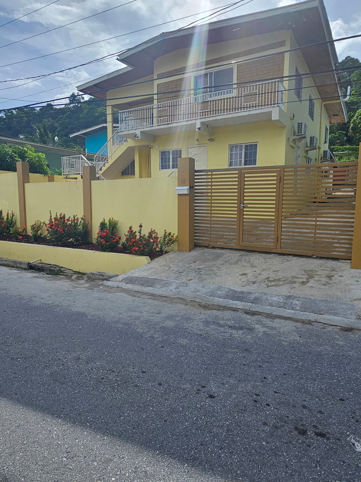 A two-story yellow building is situated behind a wooden fence with vibrant flowering plants lining the front. The entrance is marked by a gate, and several air conditioning units are visible on the exterior. The surroundings include a clear blue sky and well-maintained landscaping.
