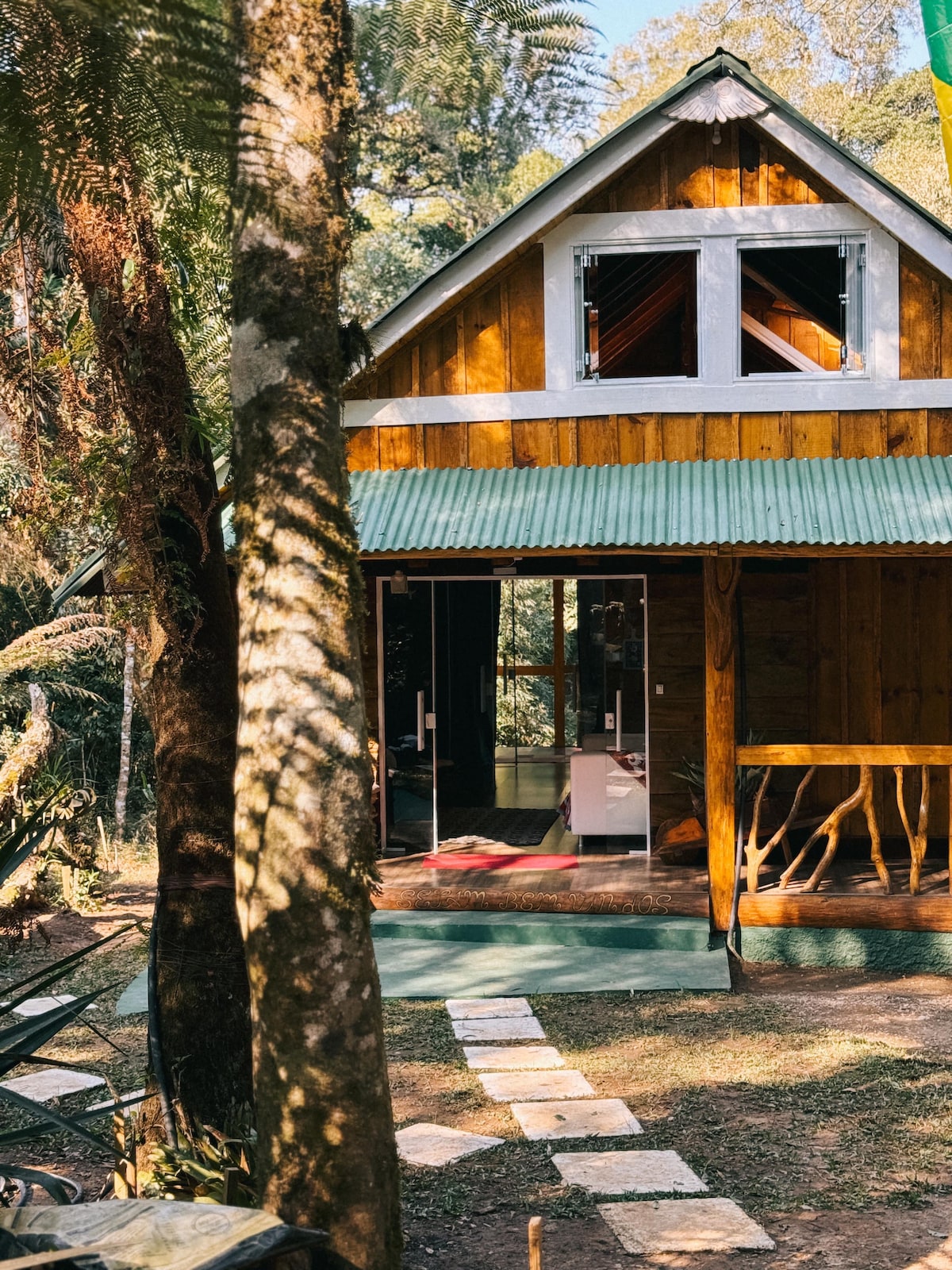 A charming cabin nestled in nature features a wooden exterior with a green metal roof. Clear double doors open to the inviting porch, surrounded by lush greenery and stone pathways that lead to the entrance, enhancing the sense of tranquility.