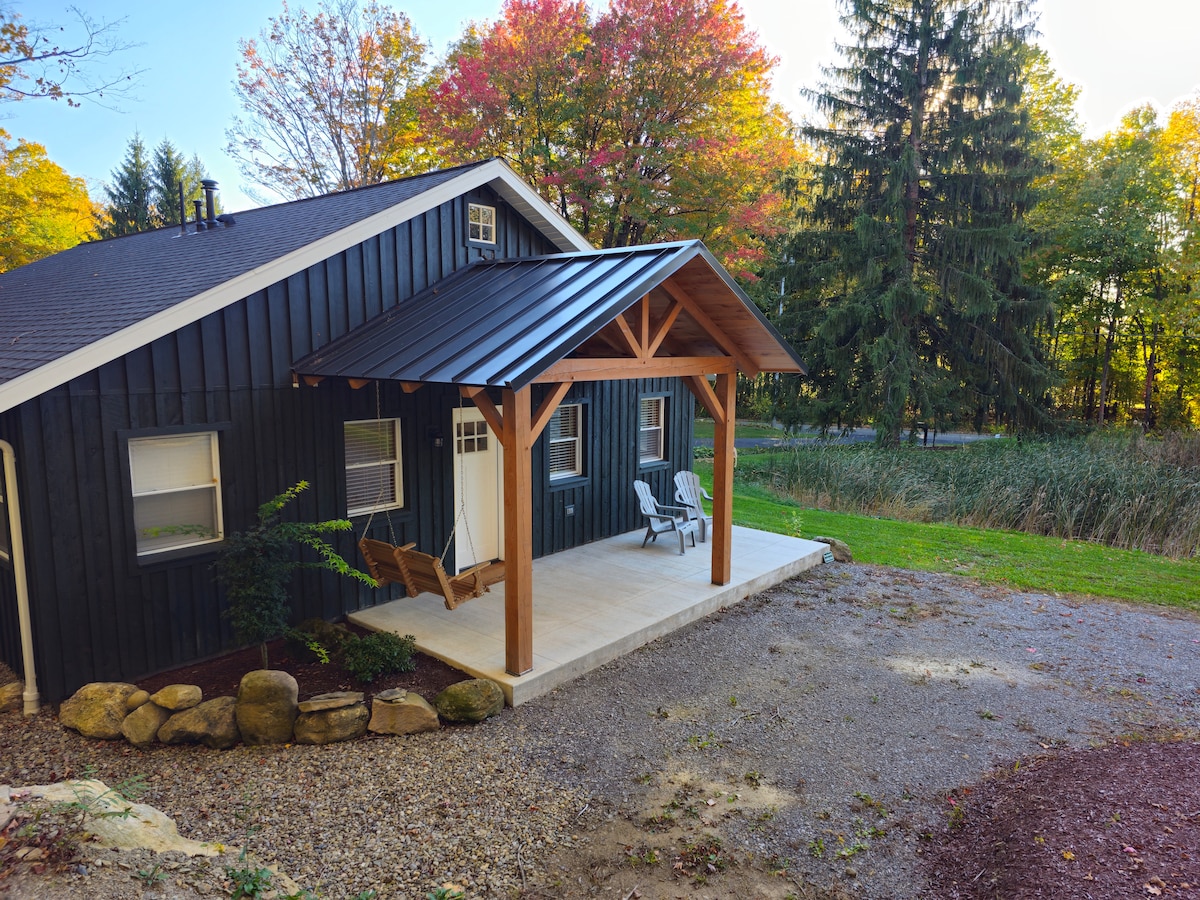 The exterior of the lodge is captured, showcasing a covered porch with a swing and two Adirondack chairs. Surrounding greenery and vibrant autumn foliage frame the scene. A gravel driveway leads to the entrance, enhancing the inviting ambiance of the lodge.