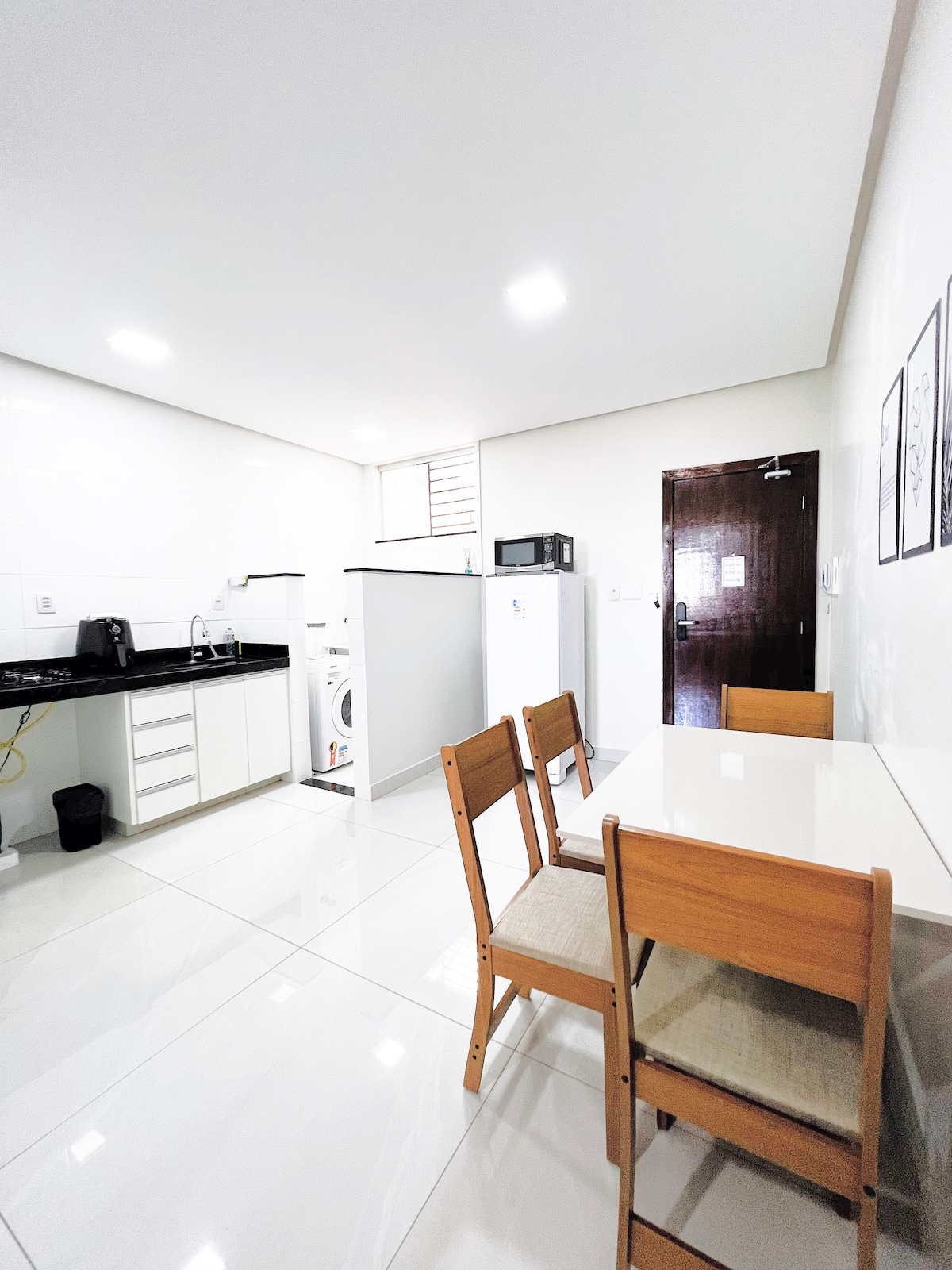 A functional kitchen and dining area is displayed, featuring a sleek black countertop and white cabinetry. A dining table with four wooden chairs is present, with a view of appliances including a refrigerator, microwave, and washing machine, all set against a clean, bright tiled floor.