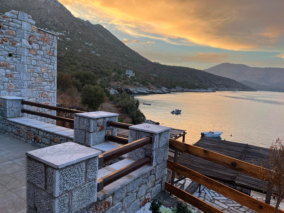 A stone terrace offers a scenic view of calm waters and distant mountains under a colorful sunset. Wooden piers extend into the water, alongside anchored boats. The warm tones of the sky reflect on the surface, creating a serene atmosphere.