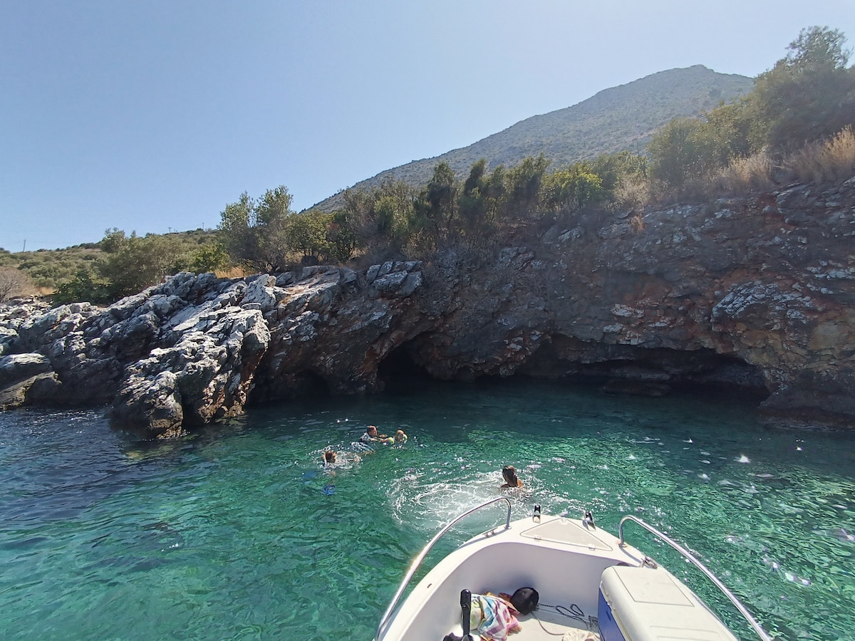 Crystal-clear turquoise waters surround a rocky coastline. A small boat is anchored nearby, with individuals swimming near an entrance to a cave. The backdrop features a mountainous landscape under a clear sky, adding to the serene coastal environment.