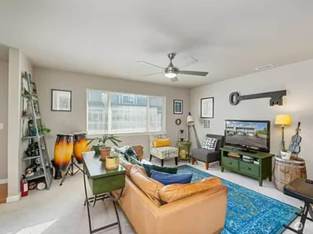 A spacious living area is filled with natural light from a large window, showcasing a comfortable sofa and eclectic furnishings. A television is positioned on a green media unit, while decorative plants and artwork enhance the cozy ambiance.