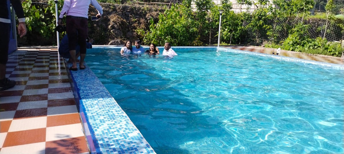 A clear swimming pool is visible, surrounded by lush greenery and a paved deck featuring colorful tiles. Several individuals are enjoying the water, with some partially submerged, while the sun reflects off the pool's surface, creating a vibrant atmosphere.