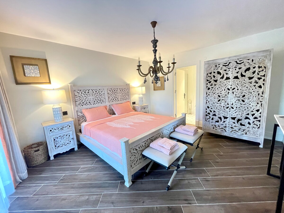 A spacious bedroom features an intricately carved wooden bed with a soft pink bedspread. Two bedside tables with lamps are positioned on either side, complemented by decorative wall art. Stylish light fixtures and a unique room divider enhance the comfortable ambiance.
