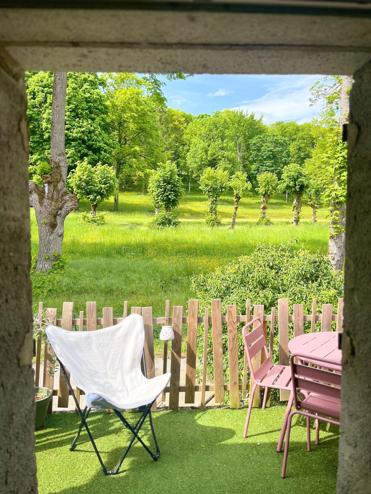A view from the doorway reveals a serene outdoor space with a rustic wooden fence. A white folding chair and a round table with three pink chairs are positioned on a grassy area, overlooking a lush green landscape filled with trees and open fields.