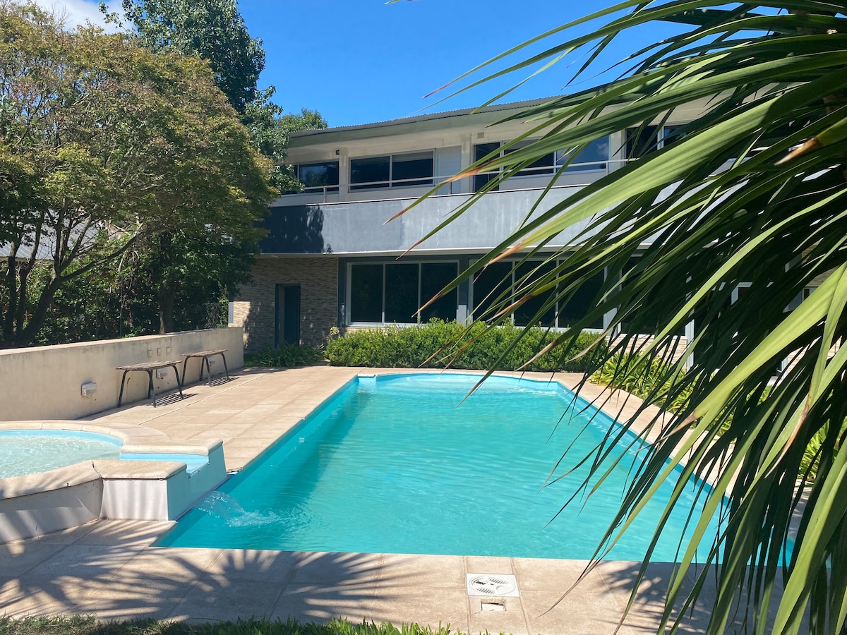 A spacious outdoor pool is framed by lush greenery and a few lounge chairs positioned nearby. The clear blue water reflects the sunlit sky, while the modern building looms in the background, enhancing the inviting atmosphere of the area.